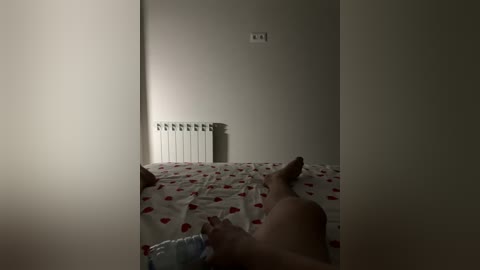 A dimly-lit video of a man lying nude on a bed with a white sheet adorned with red heart patterns. His legs are elevated, and a white radiator is visible against a plain beige wall.