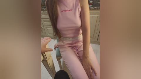 A video of a young woman with long brown hair, wearing a pink crop top and matching pants, seated on a stool in a kitchen.