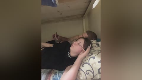 A video of a young woman with glasses and a black top, lying on a bed in a dimly lit room with a patterned comforter, looking relaxed.