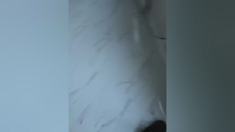 Video of a person wearing a white shirt, blurred and indistinct, standing in a dimly lit room with a pale gray wall. The image is slightly out of focus.