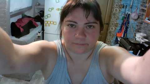 Video of a young woman with fair skin, brown hair, and a serious expression, wearing a grey tank top. Background features a cluttered room with a white door, colorful posters, and scattered clothes.
