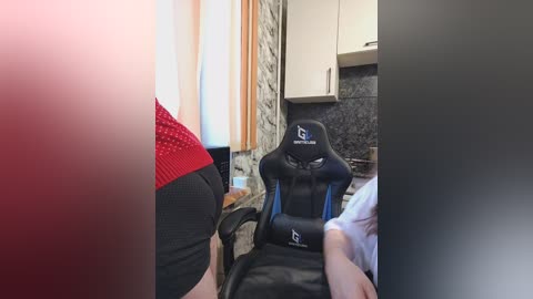 Video of a person wearing a red shirt, sitting on a black gaming chair, in a small, dimly-lit room with stone walls and a dark granite countertop.