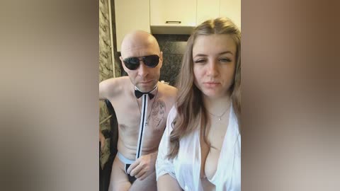 Video: A bald, muscular man in a black bow tie and briefs, standing behind a young woman with long brown hair, wearing a sheer white blouse, in a modern kitchen.