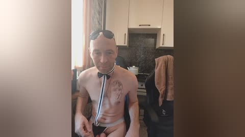 Video of a shirtless, light-skinned man with a medical stethoscope around his neck, sitting in a modern kitchen with white cabinets, black countertops, and hanging towels.