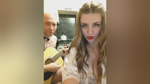 Video: A man with a bald head and a woman with long, wavy hair and red lipstick play an acoustic guitar in a modern kitchen with dark countertops.