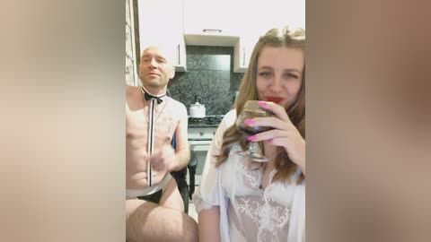 Video of a man in a white shirt and black underwear, sitting on a kitchen counter, and a woman in a white lace robe holding a glass of red wine.
