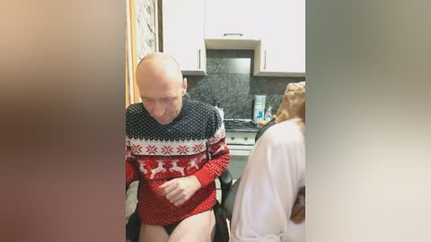 Video of a bald, fair-skinned man in a red and white Nordic sweater, sitting on a black chair in a modern kitchen with white cabinets and a black backsplash. A woman in a white robe is partially visible to the right.