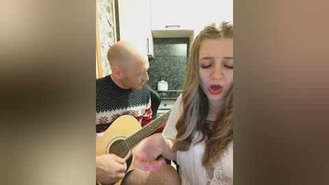 Video of a woman with long brown hair, singing passionately while strumming an acoustic guitar in a modern kitchen with white cabinets and black backsplash.