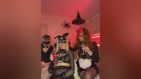 Video of three women in Halloween costumes: a blonde in black lingerie, a redhead in a witch mask, and a brunette in a maid outfit. Background features spider decorations and red lighting.