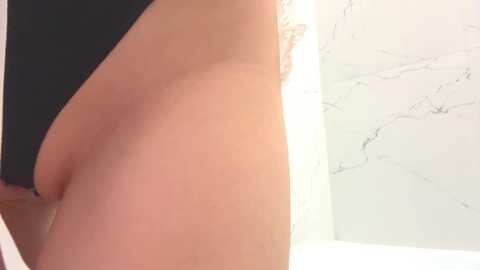 A close-up video of a fair-skinned person's buttocks in black underwear, positioned against a marble-tiled background. The image focuses on the right side of the body, emphasizing the smooth, rounded contours of the buttocks.