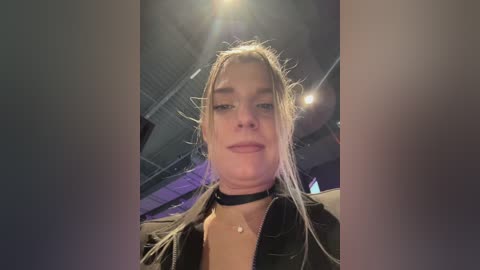 Video of a young woman with long, blonde hair, wearing a black choker and jacket, indoors with dim lighting and industrial ceiling.
