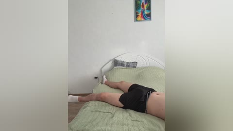 Video of a person lying on a bed in a sparsely decorated room, wearing black shorts and white socks. The room features a colorful, abstract painting on the wall and a white metal bed frame.