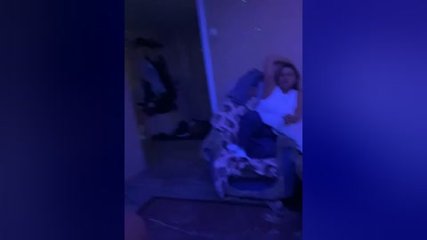 A dimly lit video shows a woman in a white shirt sitting on a chair, her head slightly bowed, with a mess of clothes and objects scattered around.