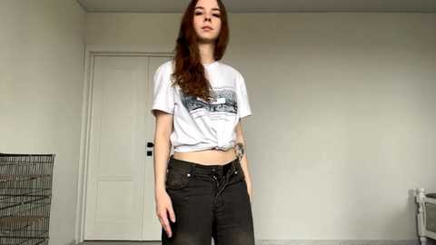 Video of a young woman with long, straight auburn hair, wearing a white graphic tee tied at the waist and dark jeans. She stands in a minimalist, white-walled room with a door and a wire mesh shelf in the background.