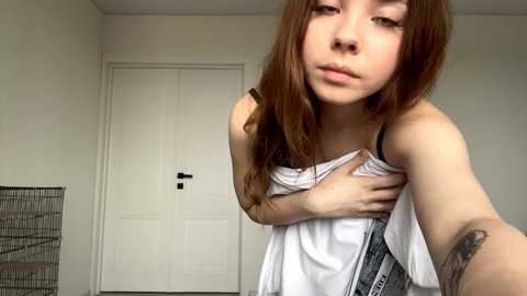 Video of a young Asian woman with long, straight brown hair, wearing a white tank top and blue jeans, standing in a minimalist, white-walled room with a closed door and a metal wire rack.