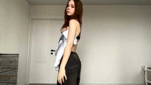 Video of a young woman with long brown hair, wearing a white tank top and tight black jeans, standing in a minimalist room with a white door, a metal rack, and a radiator.