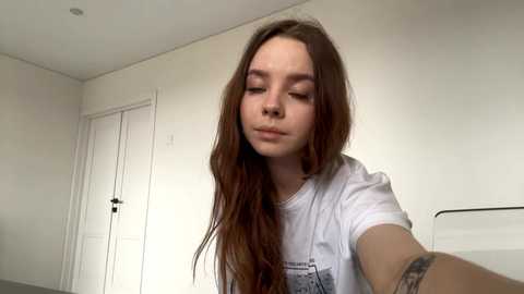 Video of a young Caucasian woman with long, wavy brown hair, wearing a white T-shirt, and a tattoo on her forearm. She has a neutral expression, standing in a simple, sparsely furnished room with white walls and a closed door.
