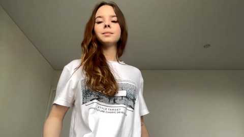 Video of a young Caucasian girl with long, wavy brown hair, wearing a white t-shirt with a black graphic design. She stands in a plain, beige-walled room with a simple, modern ceiling.