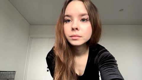 Video of a young woman with long, straight brown hair, fair skin, and blue eyes, wearing a black velvet top, standing in a simple, white-walled room with a closed door and a fan in the background.