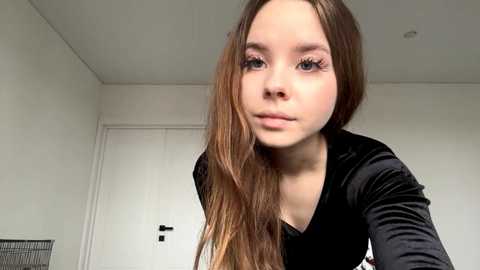 A video of a young Caucasian woman with long, light brown hair, wearing a black velvet top, in a minimalistic, white-walled room.