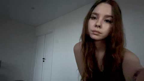 Video of a young Asian woman with long, straight, reddish-brown hair, wearing a black sleeveless top, standing in a dimly lit, minimalist white room with a closed white door and a small window.