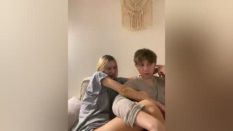 Video of a young girl with blonde hair in a grey T-shirt and shorts, comforting a boy with short brown hair in a grey T-shirt, sitting on a white couch against a beige wall with a macram\u00e9 hanging, soft lighting.