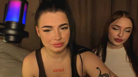 Video of two young women with pale skin and dark hair, one with a high ponytail and tattoos, the other with long hair and makeup, standing indoors, dim lighting.