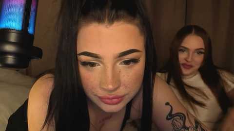 Video of a young woman with dark hair and prominent eyebrows, wearing a black top, looking down. Another woman with long hair and a tattooed arm is in the background.