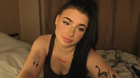 Video of a young woman with pale skin, dark hair, and tattoos, wearing a black tank top, leaning forward in a dimly-lit room with brown curtains and a white bed.