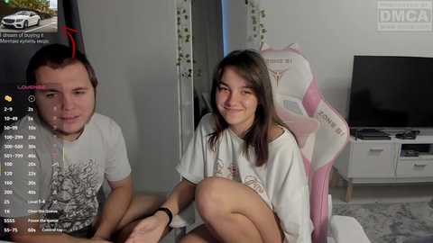 Video of a young woman with straight brown hair, wearing a white T-shirt, sitting on a pink gaming chair, smiling.