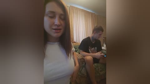 Video of a young woman with straight, brown hair, wearing a white top, smiling while partially obscured. A young man in a black T-shirt and shorts, holding a gaming controller, sits on a patterned couch in a dimly lit room with beige curtains.
