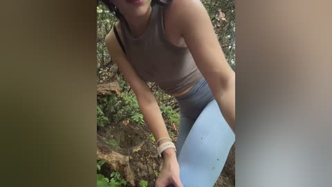A video captures a woman with medium skin tone, wearing a gray tank top and blue leggings, kneeling in a forest clearing, surrounded by greenery and fallen logs.