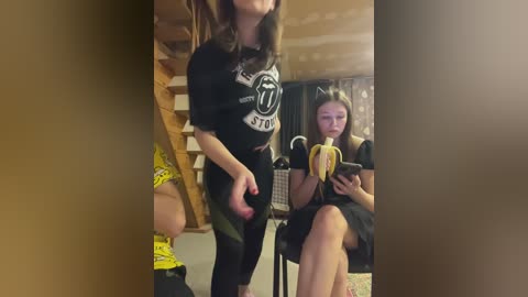 Video of three young women in a cozy, dimly lit room; one dancing, one seated with a banana, and another partially visible.