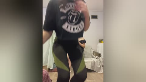 A video of a person wearing a black graphic tee and see-through black leggings with green accents, taken in a dimly lit room with a bed and a green curtain in the background.