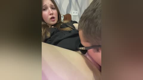 A video depicting a young girl with long brown hair in a black shirt, crying, lying on a light-colored surface, with a young boy with glasses licking her vagina.