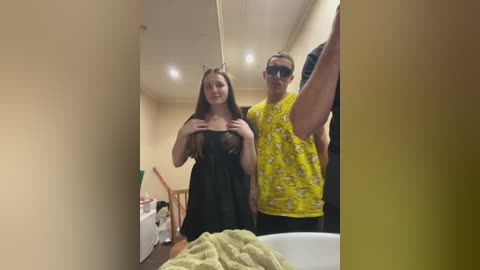 A video shows a young woman with long brown hair in a black dress, standing next to a man with sunglasses and a yellow shirt. They're in a bathroom with beige walls, a mirror, and a white sink.