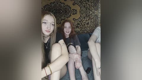Video of two young women sitting on a couch with patterned wallpaper in the background. One has long brown hair, wearing a choker, and the other has red hair, smiling.
