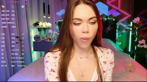 Video of a young woman with fair skin, long brown hair, and a floral dress, making a kissy face in a brightly lit room with neon lights and flowers.