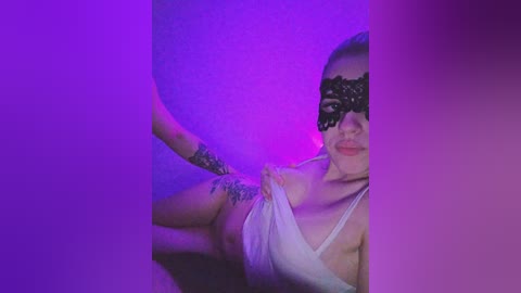 Video of a light-skinned woman with fair hair, wearing a black lace mask and a white tank top, partially removed to reveal a tattoo on her arm. The background is a vibrant blend of purple and blue hues, creating a dramatic, moody atmosphere.