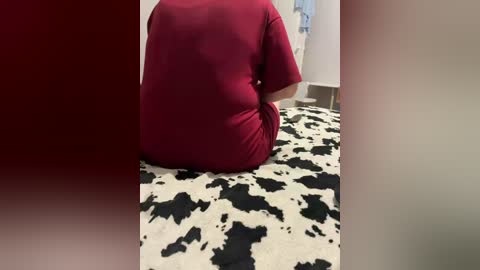 Video of a person sitting on a black-and-white cow print rug, wearing a red shirt. The background shows a white wall and a hint of a blue towel hanging.