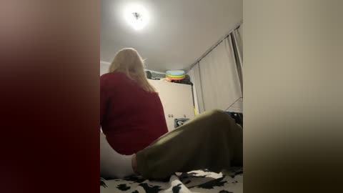 Video of a blonde woman with a large belly, wearing a red sweater and green pants, sitting on the floor in a cluttered, dimly lit room with a cow-print rug and a ceiling light.