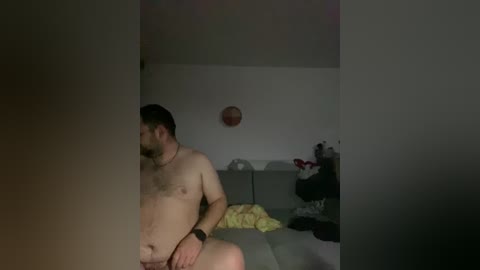 A dimly-lit video captures a nude, overweight man with a beard and short hair, sitting on a bed in a small, cluttered bedroom. The background includes a basketball, clothes, and a wall-mounted clock.