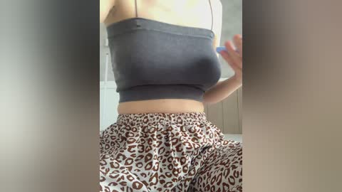 A video of a light-skinned woman with a slim physique, wearing a tight, dark gray sports bra and loose, leopard-print pajama pants, taken indoors against a blurred, neutral-toned background.