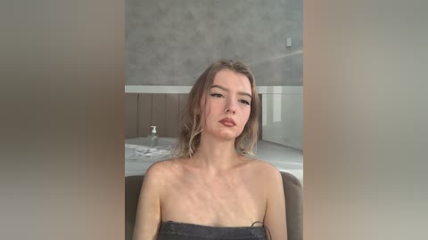 Video of a young Caucasian woman with shoulder-length, wavy, light brown hair, wearing a black towel, sitting on a chair in a modern bathroom with a white sink and a gray tiled wall.