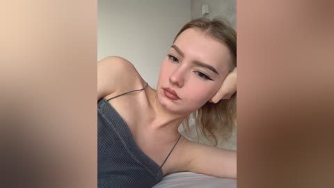 Video of a young Caucasian woman with light skin, long blonde hair, and thin eyebrows, wearing a gray off-shoulder top, lying on a bed, with a blurred background.