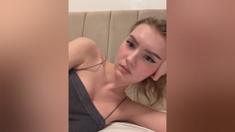 A video of a young Caucasian woman with light skin, shoulder-length blonde hair, and a slim physique, wearing a dark gray off-shoulder top, lying on a beige bed. The image is framed by vertical beige panels.