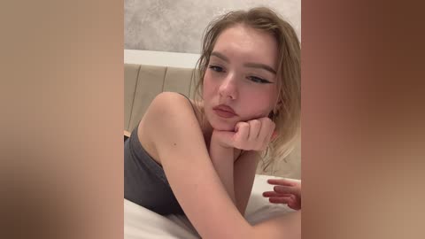 Video of a young Caucasian woman with light skin, blonde hair, and large eyes, lying on a bed, wearing a gray tank top, resting her chin on her hand, in a casual, intimate setting.