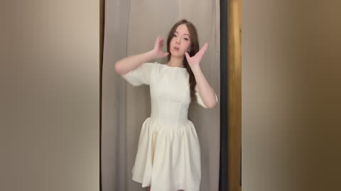 Video of a young Asian woman with light skin, long wavy brown hair, wearing a white, short-sleeved, puffy-sleeve dress, posing with hands up in front of beige curtains.