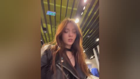A blurred video of a young woman with long, wavy brown hair, wearing a black leather jacket, standing in a modern, dimly lit indoor space with a ceiling of black and green stripes.