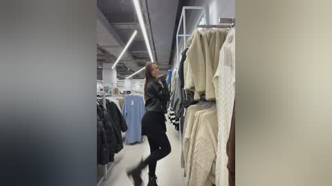 Video of a woman in a black leather jacket and pants browsing through racks of clothing in a modern, well-lit clothing store.